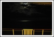 Moon hidden behind the cloud, but you can see the reflection on the water.  Ambient light from a window reflecting on the rail.